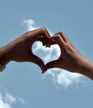 Two hands forming a heart shape against a blue sky, framing a white cloud in the background.